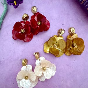 Peut inclure: Trois paires de boucles d'oreilles en forme de fleur, rouges, dorées et blanches. Chaque boucle d'oreille a un clou doré et un motif de fleur à plusieurs pétales. Les boucles d'oreilles sont sur une surface violette, avec une fleur violette et une bouteille de lavande en arrière-plan.