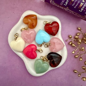 May include: A selection of heart-shaped charms in shades of brown, pink, red, and blue, presented on a white, heart-shaped dish. A perfume bottle is in the background. The charms are fitted with gold-coloured clasps.