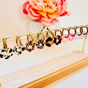 Peut inclure: Une présentation de boucles d'oreilles créoles en forme de fleur colorées avec différents motifs, dont l'imprimé léopard, la carapace de tortue et des paillettes. Les boucles d'oreilles sont suspendues à un support doré avec une base en bois.