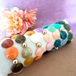 Peut inclure: Un bracelet à plusieurs rangs orné de grandes perles ovales de différentes couleurs, notamment orange, marron, rose et vert. Des perles sphériques dorées séparent les perles plus grandes. Le bracelet est présenté sur une surface blanche.