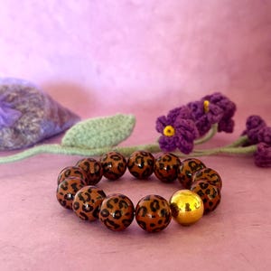 Può includere: Un braccialetto di perline con perline marroni e nere con stampa leopardata e una singola perlina dorata. Il braccialetto è esposto su una superficie rosa, con un fiore viola all'uncinetto e un sacchetto di lavanda sullo sfondo.