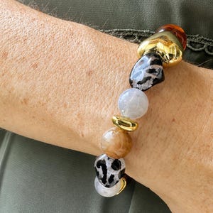 Peut inclure: Un bracelet élastique composé d'un mélange de perles, incluant des sphères lisses translucides couleur ambre, des pierres marbrées grises, des perles à motif léopard noir et blanc, et des intercalaires métalliques dorés polis disposés en séquence.