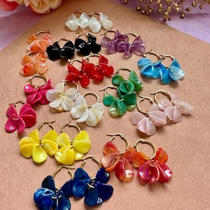 Pode incluir: Uma coleção de brincos de argola com um design floral. Os brincos apresentam argolas douradas e três pingentes em forma de pétala em várias cores, incluindo branco, preto, vermelho, roxo, azul, verde, rosa, amarelo e laranja.