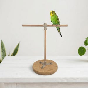 May include: A bright green and yellow budgerigar perched on a wooden T-shaped bird stand. The stand features a circular wooden base, a vertical wooden dowel, and a horizontal wooden perch connected by light-colored plastic hardware components.