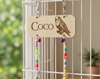 May include: A wooden pet name tag with the name "Coco" engraved, along with a detailed illustration of a parakeet. The tag hangs from a birdcage with two beaded toy strands in various colors, including pink, green, and yellow, each ending with a bell.