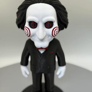 May include: A figurine of a character from the Saw horror film series. The figure has a white face with red spiral cheeks and eyes, black hair, and wears a black suit with a red bow tie. The figure is standing on a black base.