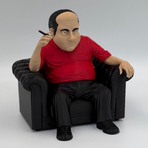 May include: A small figurine of a man sitting in a black armchair. He is wearing a red shirt, black pants, and black shoes. He has a cigarette in his mouth and a serious expression on his face.