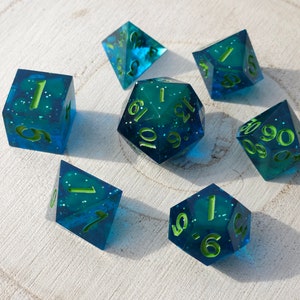 May include: Set of seven blue and green dice with white numbers. The dice are made of clear plastic with a glittery finish. The numbers are outlined in green.