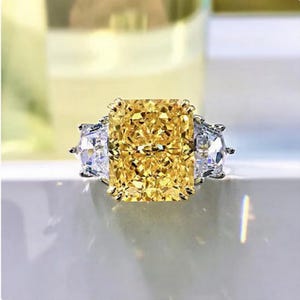 Könnte beinhalten: Ein dreisteiniger Ring mit einem großen, quadratischen, gelben Diamanten in der Mitte und zwei kleineren, runden, klaren Diamanten auf jeder Seite.