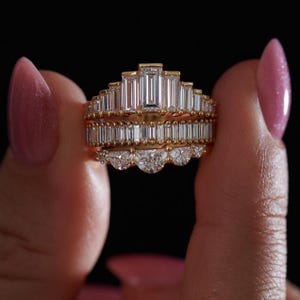 May include: A set of three gold rings with clear, sparkling gemstones. The top ring features a stepped design with rectangular stones, the middle ring has a row of smaller rectangular stones, and the bottom ring has a scalloped row of round stones. The rings are held against a black background.