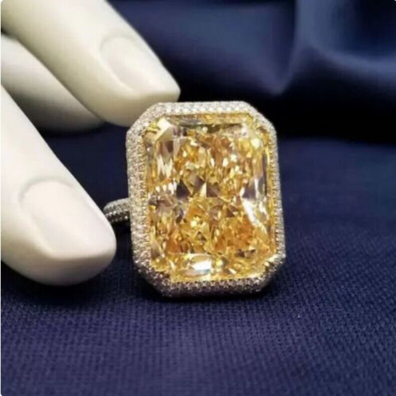 May include: A large, radiant, yellow diamond engagement ring with a square-shaped centre stone and a halo of smaller diamonds. The ring is set in a white gold band.