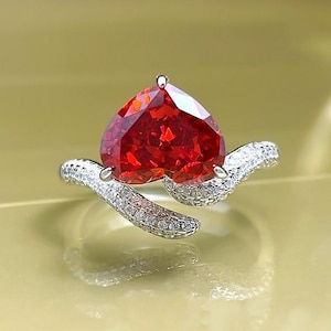 Può includere: Un anello d'argento con una grande gemma rossa sfaccettata a forma di cuore. La fascia è impreziosita da piccole pietre bianche scintillanti. L'anello è posto su uno sfondo dorato tenue, che mette in risalto il rosso vibrante della pietra centrale.
