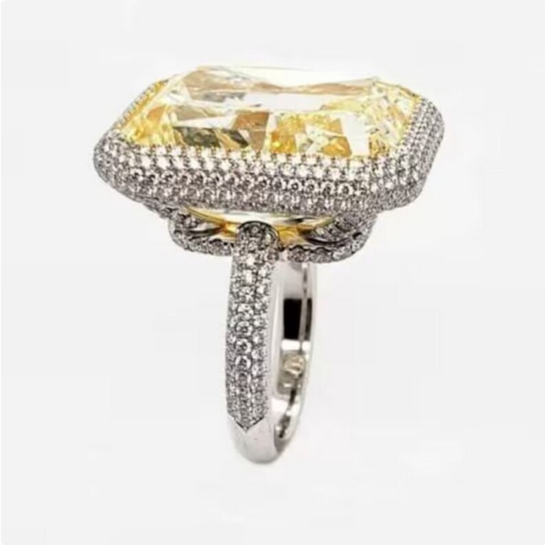 May include: A large, yellow, emerald-cut diamond ring with a halo of smaller diamonds. The ring is set in white gold and has a thick band.