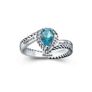 Puede incluir: Un anillo plateado con una piedra preciosa azul claro en forma de pera. El anillo presenta el nombre "Diana" y el año "2023", junto con "Western High School". La banda tiene un diseño de cuerda trenzada.