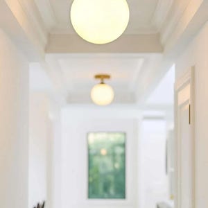May include: A bright hallway featuring two spherical, frosted glass ceiling lights with brass-coloured bases. The white walls and ceiling are complemented by the warm glow of the lights. A window at the end of the hall provides a view of the outside greenery.