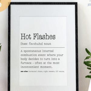 May include: A framed print with the words "Hot Flashes" in a black font. The definition of hot flashes is below the title. The print is in a black frame and is on a wooden surface.