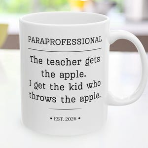 May include: A white ceramic mug featuring black text that reads: PARAPROFESSIONAL, The teacher gets the apple. I get the kid who throws the apple. EST. 2026. The mug has a standard handle and a smooth, cylindrical body with a clean, minimalist design.