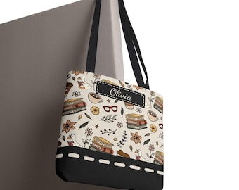 May include: A beige tote bag with a black base and handles. The bag features a pattern of books, flowers, coffee cups, and a duck reading. The name "Olivia" is displayed on a black label. The bag is hanging on a gray surface.