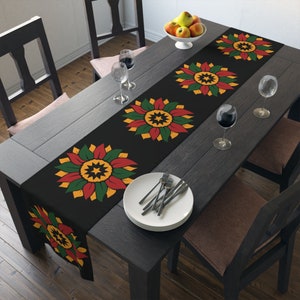 May include: Black table runner with a repeating pattern of yellow, red, and green stylized flowers. The runner is on a dark wood table with two chairs and a glass of wine.