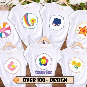 May include: Six white t-shirts with different Care Bear designs printed on the front. The designs include a rainbow, a cloud with raindrops, a star, a flower, a heart with a sunbeam, and two sunflowers. The text "OVER 100+ DESIGN" is printed at the bottom of the image.
