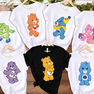 May include: Set of seven white t-shirts with different Care Bear characters printed on them. The characters include Cheer Bear, Funshine Bear, Grumpy Bear, Good Luck Bear, Wish Bear, and Friend Bear. One shirt is black with a yellow Care Bear.