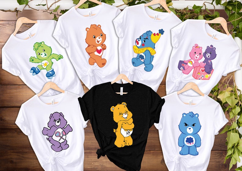May include: Set of seven white t-shirts with different Care Bear characters printed on them. The characters include Cheer Bear, Funshine Bear, Grumpy Bear, Good Luck Bear, Wish Bear, and Friend Bear. One shirt is black with a yellow Care Bear.