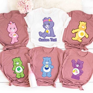 May include: Six pink t-shirts with different Care Bear characters printed on them. The characters include Cheer Bear, Funshine Bear, Good Luck Bear, Grumpy Bear, Love-A-Lot Bear, and Tenderheart Bear. The shirts have the text "Custom Text" printed on one of the shirts.