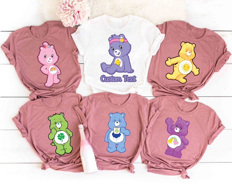 May include: Six pink t-shirts with different Care Bear characters printed on them. The characters include Cheer Bear, Funshine Bear, Good Luck Bear, Grumpy Bear, Love-A-Lot Bear, and Tenderheart Bear. The shirts have the text "Custom Text" printed on one of the shirts.