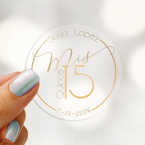 May include: A circular clear sticker featuring gold foil text that reads Sofia Lopez, Mis Quince 15, and the date 7-18-2026. The design includes a thin gold circular border. The sticker is held up against a blurred background.