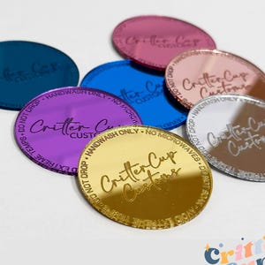 May include: A collection of colorful, circular acrylic cup toppers in various shades, including teal, pink, blue, purple, gold, rose gold, and silver. Each topper features the text "Critter Cup Customs" and care instructions.