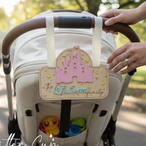 May include: A wooden stroller tag featuring a pink castle design and the text "The Williams Family." The tag is attached to a cream-colored stroller. The stroller has a brown handle and white straps. The tag is decorated with yellow stars.