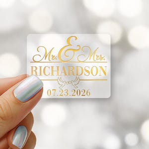 May include: A clear rectangular sticker with gold foil text. The text reads "Mr. & Mrs. RICHARDSON" and "07.23.2026". The sticker has a decorative design of two doves. The logo "Critter Cup customs" is in the upper left corner.