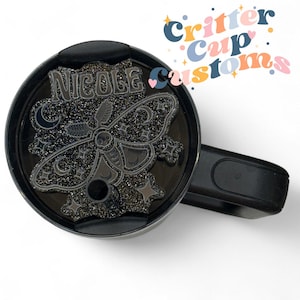 May include: Black tumbler lid with a decorative, glittery moth design. The moth has a skull on its body and is surrounded by moons and stars. The name "Nicole" is above the moth. The tumbler lid has a handle and a drinking hole.