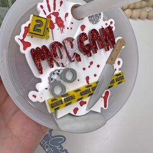 Może przedstawiać: Biała plastikowa pokrywka kubka z motywem miejsca zbrodni. Pokrywka ma czerwone plamy krwi, ż&oacute;łtą taśmę z napisem &bdquo;miejsce zbrodni&rdquo;, n&oacute;ż, kajdanki, odcisk palca i imię &bdquo;MORGAN&rdquo; w czerwonych literach z brokatem. Liczba &bdquo;2&rdquo; znajduje się na ż&oacute;łtym kwadracie.