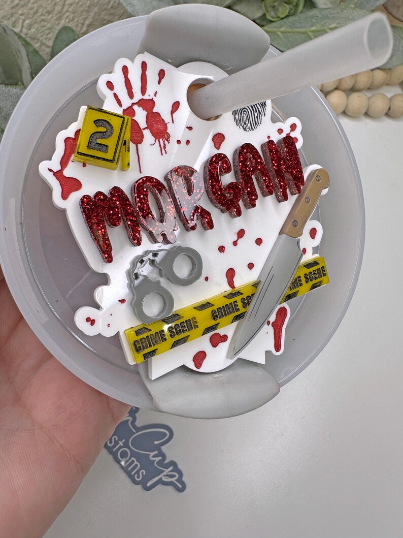 Może przedstawiać: Biała plastikowa pokrywka kubka z motywem miejsca zbrodni. Pokrywka ma czerwone plamy krwi, ż&oacute;łtą taśmę z napisem &bdquo;miejsce zbrodni&rdquo;, n&oacute;ż, kajdanki, odcisk palca i imię &bdquo;MORGAN&rdquo; w czerwonych literach z brokatem. Liczba &bdquo;2&rdquo; znajduje się na ż&oacute;łtym kwadracie.