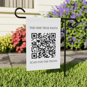 Op de afbeelding: Een witte tuin vlag met zwarte tekst die "THE ONE TRUE FAITH" leest boven een QR-code en "SCAN FOR THE TRUTH" eronder. De vlag wordt getoond op een zwarte metalen standaard in een tuin met bloemen.