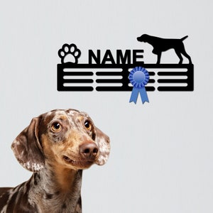 May include: Black metal dog award rack with a paw print, the word "NAME", and a blue ribbon. A brown and white spotted dog looks up at the rack.
