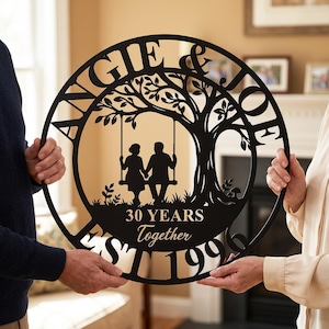 Peut inclure: Une enseigne murale circulaire en métal noir avec une découpe représentant un arbre et un couple assis sur une balançoire. Le texte indique ANGIE & JOE, 30 YEARS Together et EST 1996. Le motif est encadré par un bord circulaire noir.