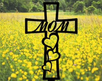 May include: Black metal cross with the word "MOM" on the crossbar and three heart shapes hanging from the bottom. The cross is in front of a field of yellow flowers.