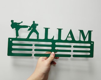 Könnte beinhalten: Grünfarbener Medaillenhalter aus Metall mit 10 Fächern für Medaillen. Der Halter zeigt zwei Karate-Figuren und den Namen "LIAM" in großen Buchstaben.