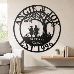 Puede incluir: Un letrero de pared circular de metal negro con la silueta de una pareja en un columpio bajo un &aacute;rbol. El texto dice ANGIE & JOE, 30 YEARS Together y EST 1996. El dise&ntilde;o est&aacute; cortado de una sola pieza de metal con un acabado negro mate.