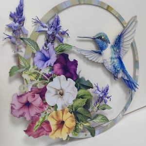 May include: A blue hummingbird in flight with its wings spread wide, surrounded by a circle of purple, white, yellow, and pink flowers. The flowers are arranged around a circular wooden frame with a light blue and green painted design.