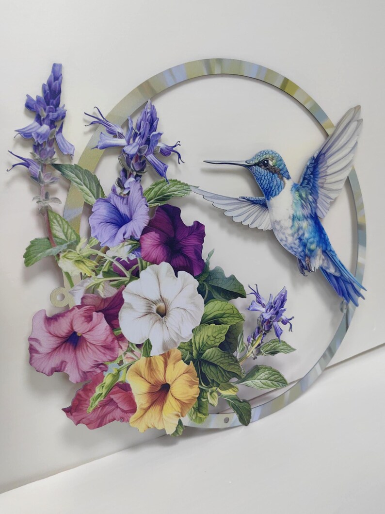 May include: A blue hummingbird in flight with its wings spread wide, surrounded by a circle of purple, white, yellow, and pink flowers. The flowers are arranged around a circular wooden frame with a light blue and green painted design.