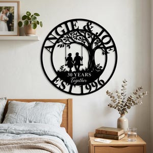 Puede incluir: Un letrero de pared circular de metal negro con la silueta de dos personas sentadas en un columpio bajo un &aacute;rbol. El texto dice ANGIE & JOE, 30 YEARS Together y EST 1996. El dise&ntilde;o est&aacute; cortado de una sola pieza de metal con acabado liso.