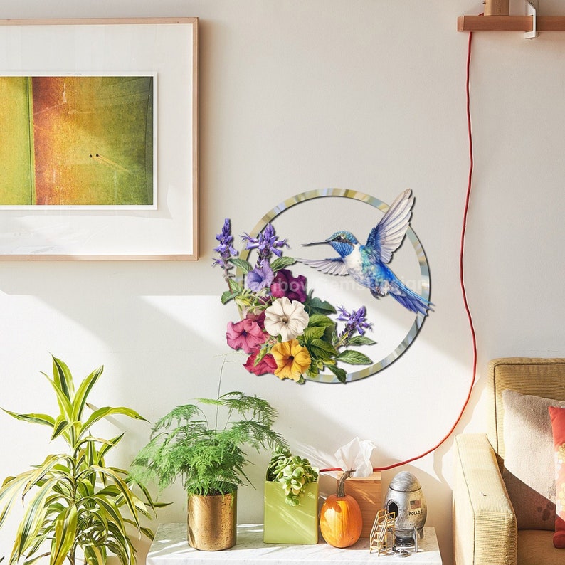 May include: A blue hummingbird with a long beak flies towards a circle of colorful flowers. The flowers are pink, purple, white, and yellow. The background is a white wall.