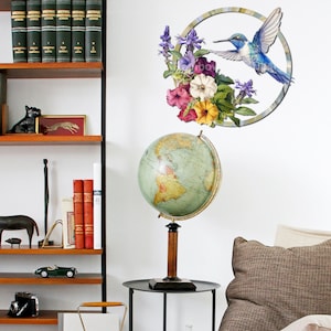 May include: A vintage globe with a wooden stand sits on a black table next to a brown couch. A wall decal with a hummingbird and flowers is on the wall behind the globe.