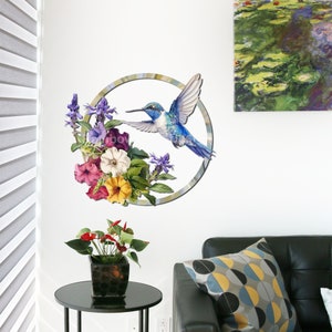 May include: A blue hummingbird with a long beak flies towards a circle of colorful flowers. The flowers are pink, purple, yellow, and white. The background is a white wall.