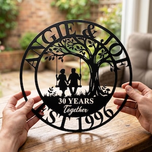 Puede incluir: Un letrero de pared circular de metal negro con la silueta de una pareja sentada en un columpio de &aacute;rbol. El texto dice ANGIE & JOE, 30 YEARS Together y EST 1996. El dise&ntilde;o incluye un gran &aacute;rbol con ramas y hojas dentro del marco circular.