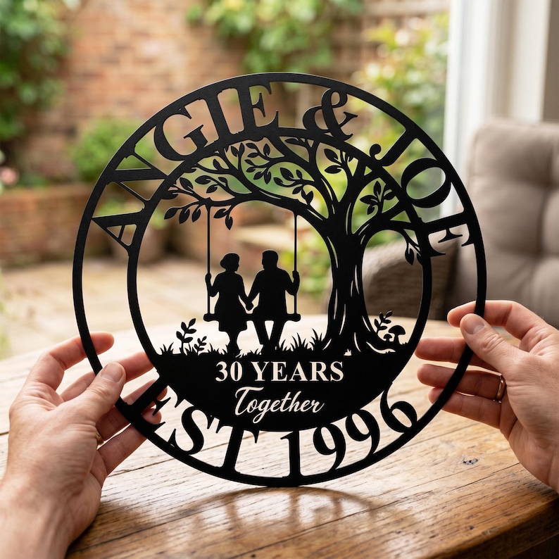 Puede incluir: Un letrero de pared circular de metal negro con la silueta de una pareja sentada en un columpio de &aacute;rbol. El texto dice ANGIE & JOE, 30 YEARS Together y EST 1996. El dise&ntilde;o incluye un gran &aacute;rbol con ramas y hojas dentro del marco circular.