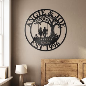 Puede incluir: Un letrero de pared circular de metal negro con la silueta de un &aacute;rbol y una pareja sentada en un columpio. El texto dice ANGIE & JOE, 30 YEARS Together y EST 1996. El dise&ntilde;o est&aacute; cortado de una sola pieza de metal con acabado negro mate.